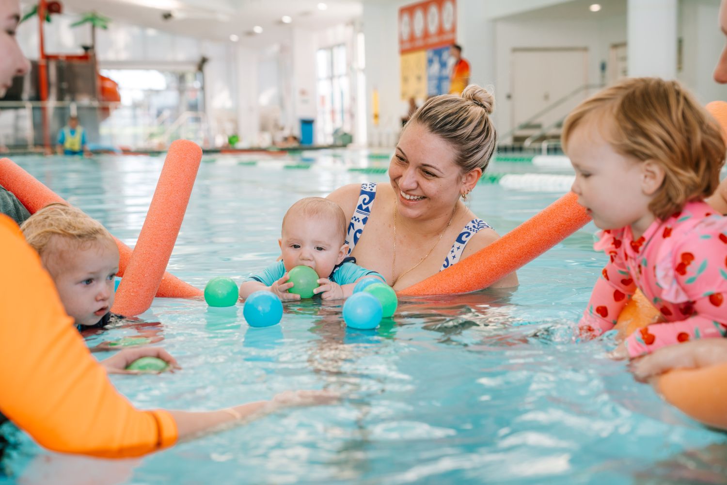 A smiling mother holds her baby boy, who is biting a green plastic ball, during an indoor pool lesson with other parents and toddlers using pool noodles.